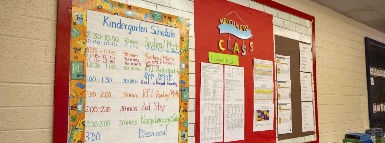 A kindergarten classroom wall features a daily schedule and a red bulletin board displaying lesson plans and classroom documents.