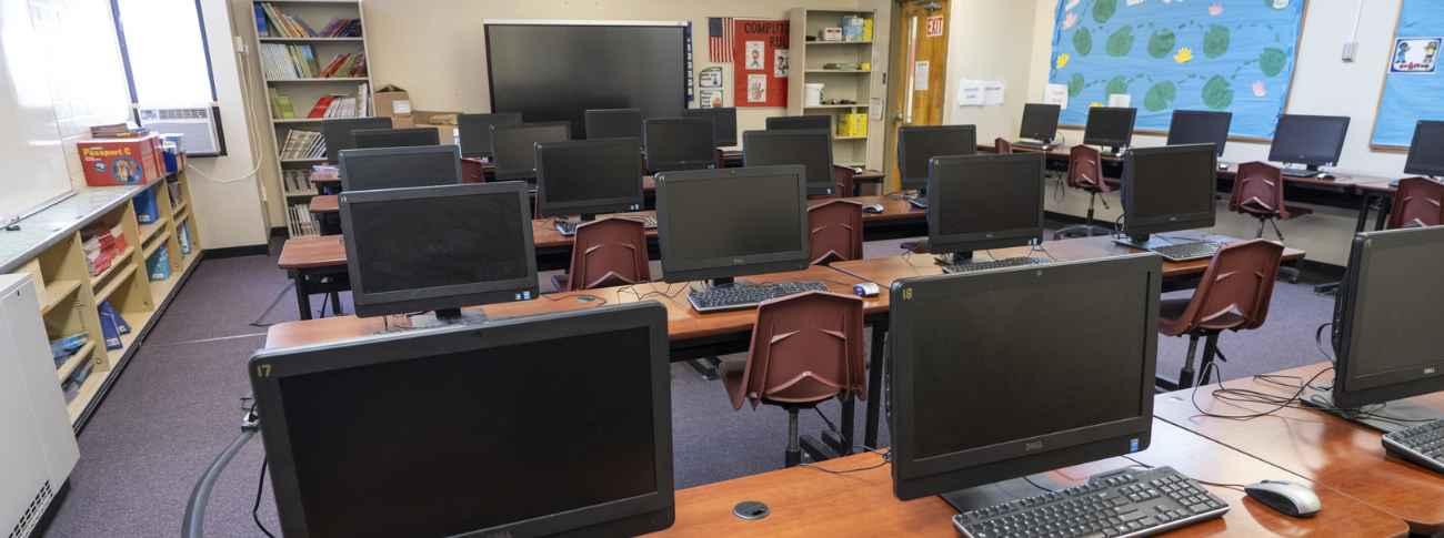 A computer lab classroom with rows of desktop computers, keyboards, and chairs arranged on tables, with bookshelves, a large display screen, and educational bulletin boards along the walls.